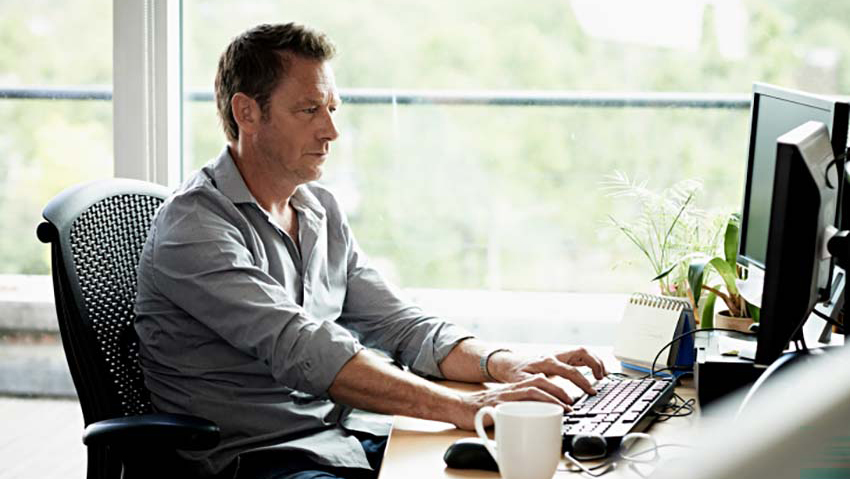 Man types on a keyboard at a desk with large windows behind him that look out over trees